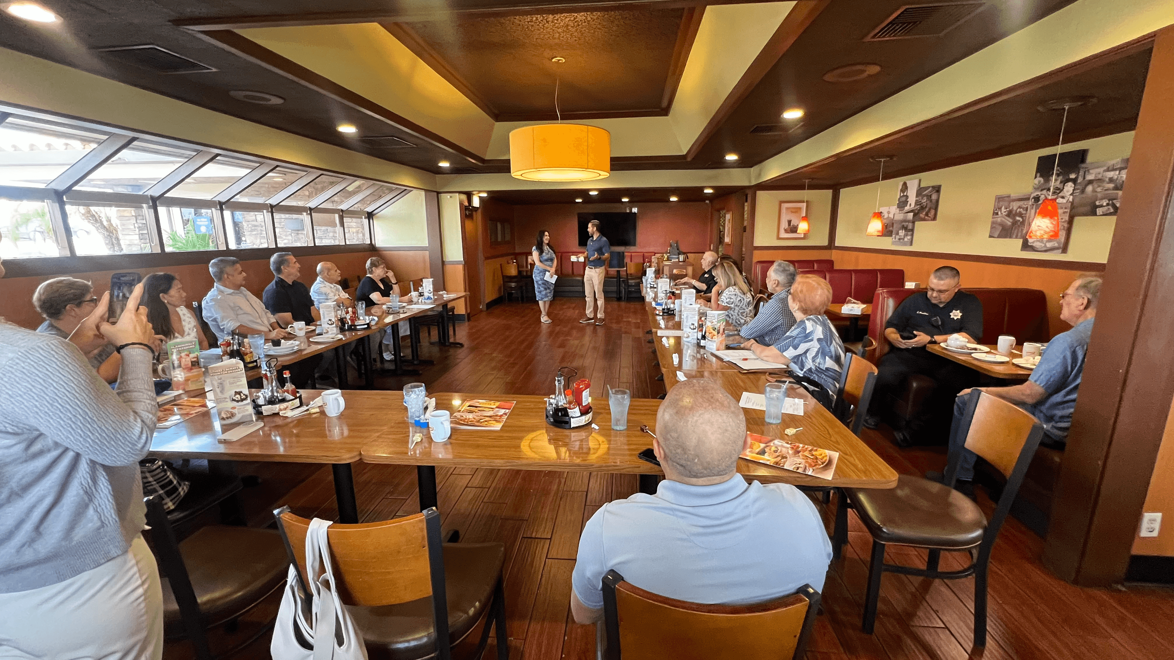 People seated around tables in a restaurant listening to a presentation by two standing speakers.