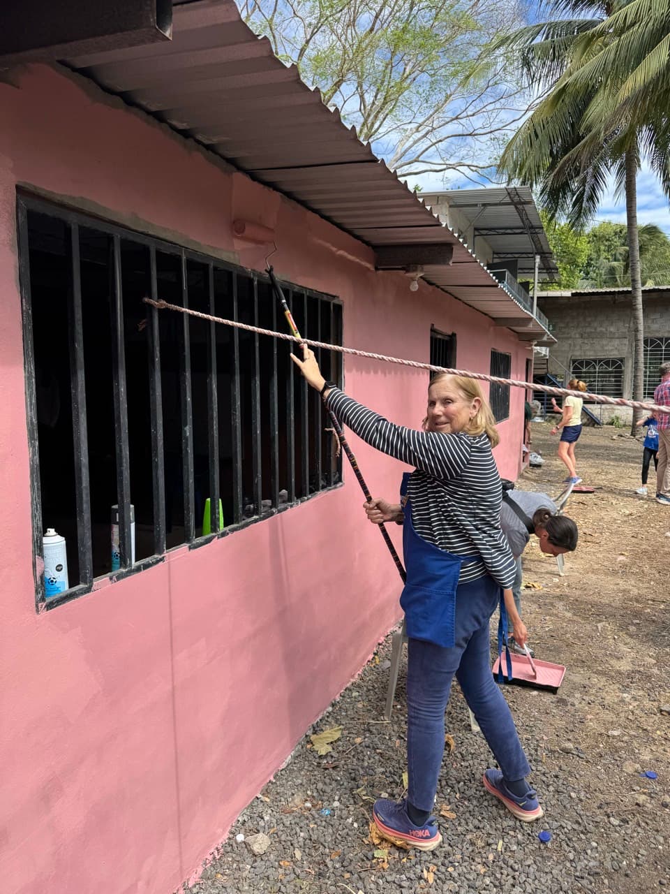 A woman in a striped shirt and blue apron uses a long-handled roller to paint a pink wall.