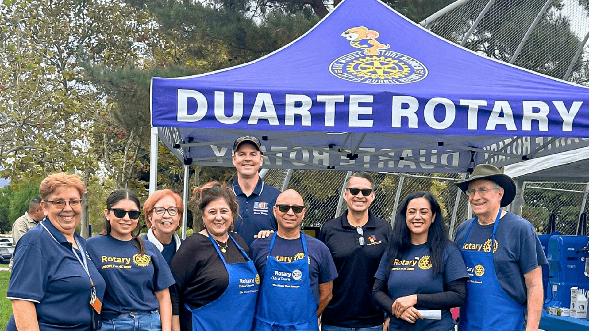 Smiling Duarte Rotary Club members in branded shirts and aprons pose under a blue tent.